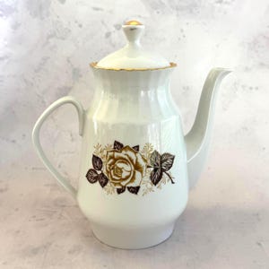 May include: White ceramic coffee pot with a lid and handle. The pot features a gold and brown rose design on the front and gold trim. The spout is curved, and the lid has a small gold finial. A vintage kitchenware item.