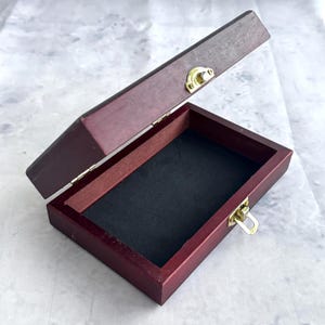 Può includere: Una scatola rettangolare in legno con coperchio incernierato, aperta per rivelare un interno nero. La scatola è tinta di un colore marrone rossastro scuro e presenta una chiusura e cerniere color oro.
