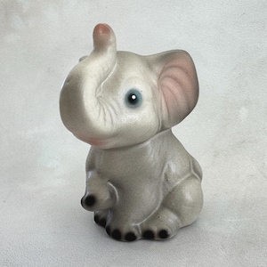 May include: A gray elephant figurine with a trunk raised upwards. The elephant has pink inner ears, blue eyes, and black toenails. The figurine is made of a smooth material and is sitting down.