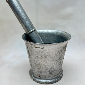 Puede incluir: Un mortero y un pilón de metal plateado. El mortero es un recipiente en forma de cuenco con un borde ligeramente acampanado. El pilón es una varilla cilíndrica con un extremo redondeado.