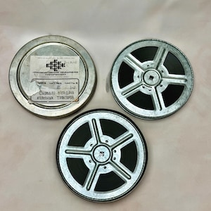 May include: Two silver metal film reels with black film strips and one round metal film canister lid. The lid features a white paper label with black Cyrillic text and a logo. The text reads "ПРОИЗВОДСТВЕННОЕ ОБЪЕДИНЕНИЕ КОПИРФИЛЬМ" and "СИЛОВАЯ ПЕРЕДАЧА КОЛЕСНЫХ ТРАКТОРОВ".
