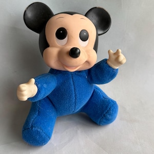 Op de afbeelding: Een blauw pluche Mickey Mouse speelgoed dat een blauwe jumpsuit draagt. Het speelgoed heeft zwarte oren en een zwarte neus. Het speelgoed heeft grote ogen en een glimlachende mond.