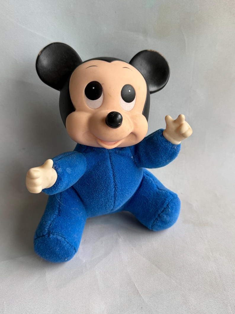 Op de afbeelding: Een blauw pluche Mickey Mouse speelgoed dat een blauwe jumpsuit draagt. Het speelgoed heeft zwarte oren en een zwarte neus. Het speelgoed heeft grote ogen en een glimlachende mond.