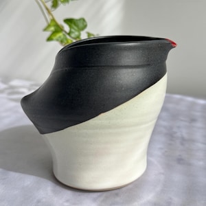 Peut inclure: Un pichet en céramique présentant un design bicolore avec une partie supérieure noir mat et un corps inférieur de couleur crème. Le récipient possède un bord large et évasé avec un petit accent rouge au bec et un élément latéral saillant.