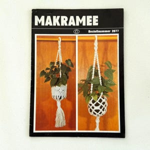 Peut inclure: Un livre vintage intitulé "Makramee" avec le numéro de commande 2077. La couverture présente deux photos de suspensions de plantes en macramé avec un cordon blanc et des plantes vertes.