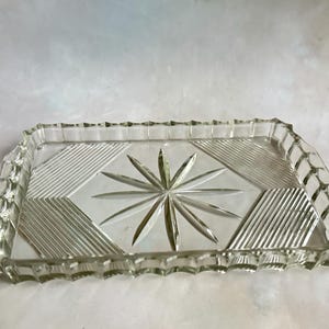Puede incluir: Una bandeja de servir de vidrio transparente con un borde festoneado y un diseño de estrella en el centro. La bandeja tiene un patrón de diamante alrededor de la estrella.