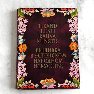 Może przedstawiać: Bordowa okładka książki ze złotymi literami i haftem kwiatowym. Tekst brzmi "TIKAND EESTI RAHVA-KUNSTIS" i "ВЫШИВКА В ЭСТОНСКОМ НАРОДНОМ ИСКУССТВЕ". Haft przedstawia pomarańczowe, różowe i zielone kwiaty i liście.