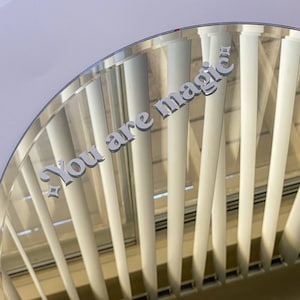 May include: A clear, semi-circular mirror with the words "You are magic" in a light gray, stylized font. A small star graphic is next to the word "magic". The mirror reflects a view of white vertical blinds.