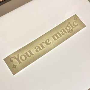 May include: A rectangular sticker with the words "You are magic" in a serif font. The text is a light brown color with a glittery effect. The sticker has a grid pattern on the surface.