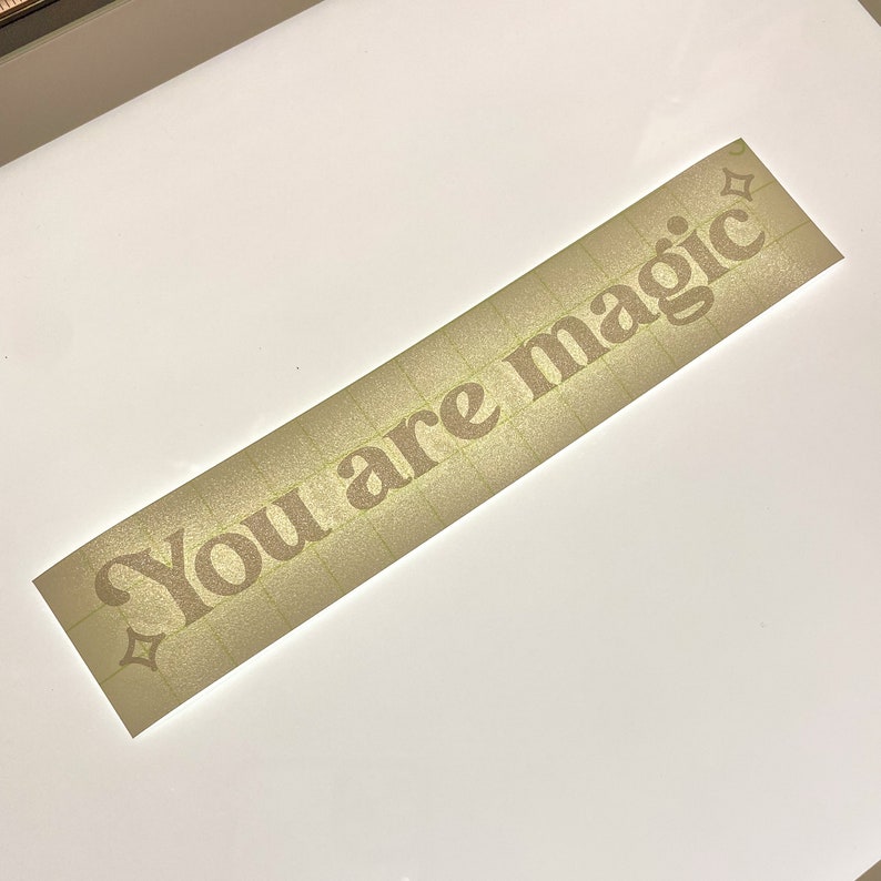 May include: A rectangular sticker with the words "You are magic" in a serif font. The text is a light brown color with a glittery effect. The sticker has a grid pattern on the surface.