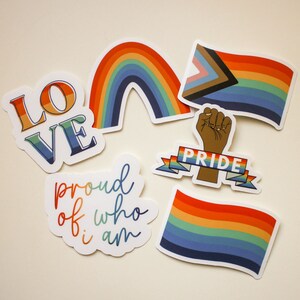 May include: A collection of colorful stickers with Pride-themed designs. The stickers include a rainbow, flags, the word "LOVE", a raised fist with a banner that says "PRIDE", and the phrase "proud of who i am".