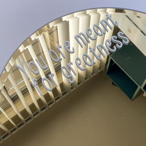 May include: A decorative, curved architectural element with a repeating vertical pattern. The text "You are meant for greatness" is displayed in a light gray, 3D font across the top of the structure. The background is a light beige color.