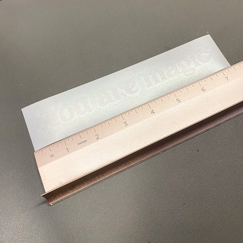 May include: A rectangular, white sticker with the words "You are magic" printed on it. A wooden ruler is placed diagonally across the sticker, measuring its length in inches. The sticker is on a gray surface.