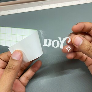 May include: A clear vinyl sticker with the word "you" in white, with a star-shaped design. The sticker is being peeled from a backing sheet with a grid pattern. The background is a light gray surface.