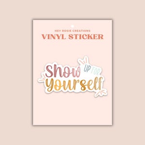May include: A vinyl sticker on a light pink backing card labeled Hey Rosie Creations. The sticker features the text Show Up For Yourself in a gradient of pink, orange, and yellow cursive lettering with small decorative lines around the words.
