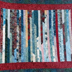 K&ouml;nnte beinhalten: Eine Decke mit rotem Rand und einem Muster aus vertikalen Streifen in Blaut&ouml;nen, Gr&uuml;n- und Rosat&ouml;nen.