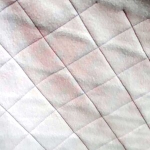 以下が含まれることがあります： ダイヤモンドのパターンが施された白いキルティング生地。生地は薄いピンクの色合いをしています。