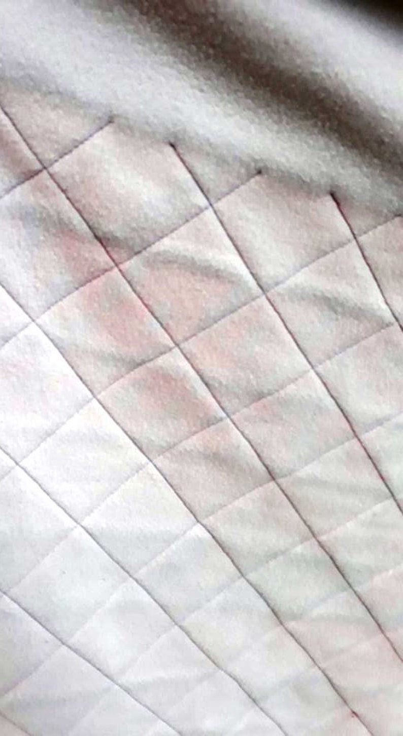 以下が含まれることがあります： ダイヤモンドのパターンが施された白いキルティング生地。生地は薄いピンクの色合いをしています。