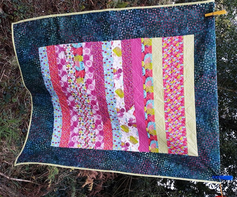 以下が含まれることがあります： パッチワークのデザインが施されたカラフルなキルト。キルトは、ピンク、ブルー、グリーン、イエローの様々な色合いの布のストリップでできています。キルトは、白いドットの入った濃い青色の布で縁取られています。