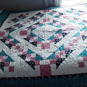 May include: A white quilt with a geometric pattern of pink, teal, and black squares and triangles. The quilt is quilted with a swirling pattern.