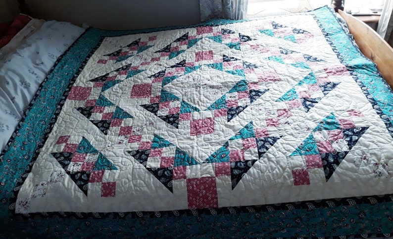 May include: A white quilt with a geometric pattern of pink, teal, and black squares and triangles. The quilt is quilted with a swirling pattern.