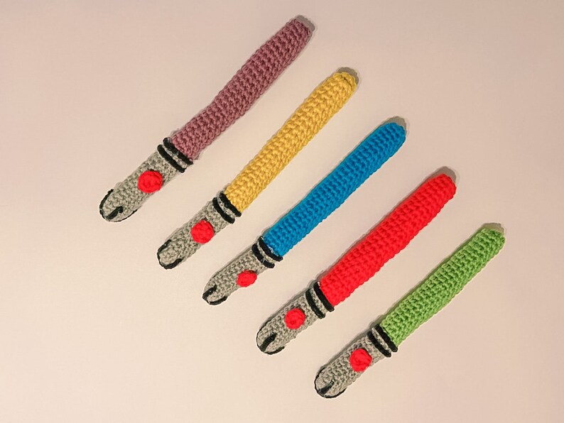 K&ouml;nnte beinhalten: F&uuml;nf geh&auml;kelte Lichtschwerter in verschiedenen Farben: Lila, Gelb, Blau, Rot und Gr&uuml;n. Jedes hat einen grauen Griff mit schwarzen Details und einem roten Knopf. Die Lichtschwerter sind diagonal auf einer wei&szlig;en Oberfl&auml;che angeordnet.