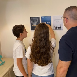 May include: Three people, two children and an adult, are looking at movie posters on a white wall. The posters are for the movies "9 East Street", "Eight Days", and "Just the Two of Us".