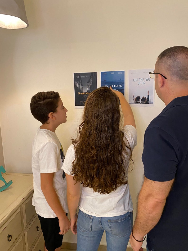 May include: Three people, two children and an adult, are looking at movie posters on a white wall. The posters are for the movies "9 East Street", "Eight Days", and "Just the Two of Us".