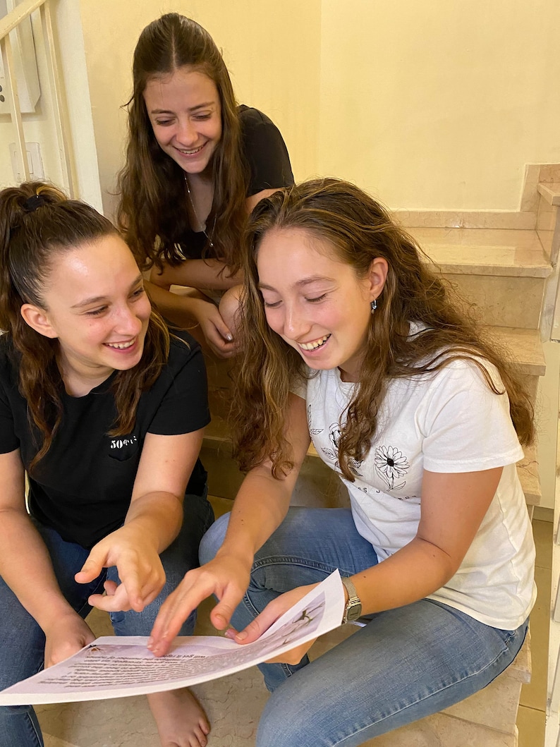 May include: Three young people are sitting on a staircase, looking at a piece of paper. They are smiling and laughing. The paper has text on it, and the person in the center is pointing at it.