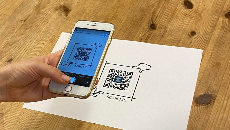 May include: A smartphone displaying a QR code with the text "Scan Me" on the screen. The phone is being used to scan a printed QR code with the text "Scan Me" on a white piece of paper.