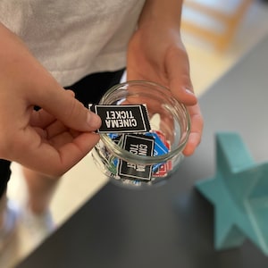 May include: A glass jar filled with small black and blue paper tickets that say "Cinema Ticket" on them. A hand is holding one of the tickets.