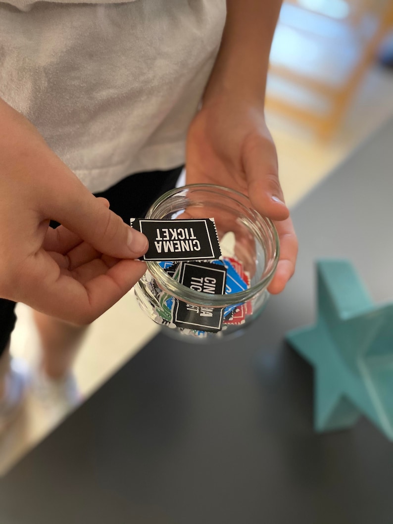 May include: A glass jar filled with small black and blue paper tickets that say "Cinema Ticket" on them. A hand is holding one of the tickets.