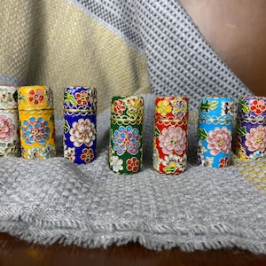 May include: Seven decorative, cylindrical containers with floral designs. Each container has a different colour scheme, including white, yellow, blue, green, red, and purple, with gold accents. The containers are arranged on a grey and yellow patterned surface.