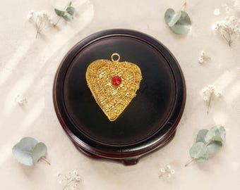 Puede incluir: Un colgante en forma de corazón de color dorado con un intrincado trabajo de metal en filigrana y una pequeña piedra roja engastada en el centro. El colgante incluye una pequeña anilla circular en la parte superior y una superficie texturizada.