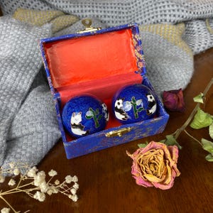 May include: A pair of blue decorative Chinese health balls with panda designs, resting in a blue box with a red interior. The box is open, revealing the balls. The background includes a gray blanket and dried flowers.