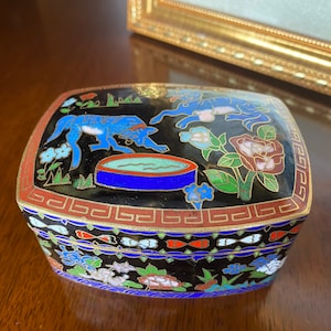 May include: A small, rectangular, black enamel box with a gold trim. The lid features a blue and white floral design with two blue animals and a blue and green bowl. The bottom of the box has a similar design.