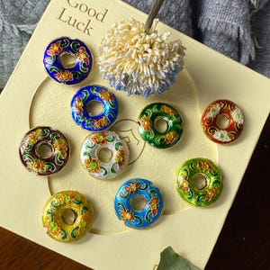 Peut inclure: Collection de perles rondes en émail coloré avec des motifs floraux. Les perles présentent une variété de couleurs, dont le bleu, le vert, le jaune et le rouge, avec des accents métalliques dorés. Les perles sont disposées sur une surface crème avec les mots "Good Luck".