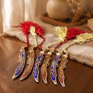 May include: A set of six decorative miniature curved swords with metallic gold handles and blades featuring colorful enamel dragon motifs. Each sword is adorned with a decorative red and cream-colored tassel attached to the hilt by a cord.
