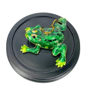 Könnte beinhalten: Eine grüne und goldene Froschfigur mit schwarzen Flecken, die auf einem schwarzen runden Sockel steht. Der Frosch hat eine goldene Schlaufe auf dem Rücken zum Aufhängen.