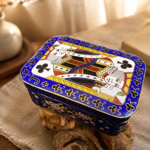 Puede incluir: Una caja metálica rectangular con un diseño de sota de tréboles en la tapa. La caja tiene un borde azul intenso decorado con símbolos de trébol dorados y la letra J. Los laterales de la caja muestran un patrón floral sobre un fondo azul.