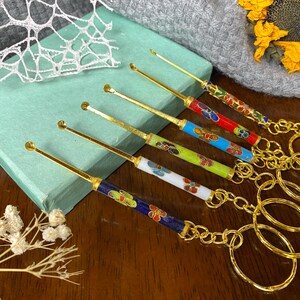 May include: A selection of decorative ear picks with colourful floral designs. Each pick has a gold-coloured handle and a matching keychain. The picks are arranged on a wooden surface next to a teal box and dried flowers.