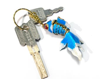 May include: A gold key chain with a blue and white enamel fish charm. The fish has a red dot on its head and gold accents. The keychain is attached to two silver keys.
