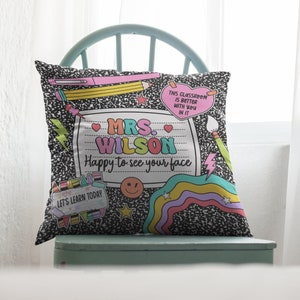 May include: A decorative throw pillow with a notebook paper design. The pillow features the text "MRS. WILSON Happy to see your face" and other school-themed graphics. The background is a black and white composition notebook pattern.