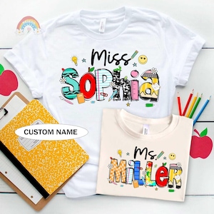 May include: White t-shirt with the name "Miss Sophia" in colorful, school-themed lettering. A second, cream-colored shirt displays "Ms. Miller". A yellow composition notebook and pencils are also visible.