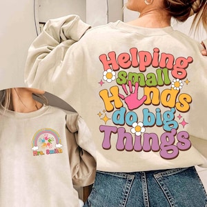 May include: A beige sweatshirt with the text "Helping small hands do big things" in colorful letters. The text is surrounded by small flowers and stars. The sweatshirt has a crew neck and long sleeves. A second image shows a beige sweatshirt with a rainbow and handprints with the text "Mrs. Smith" underneath.