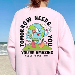 May include: Pink sweatshirt with a graphic of a smiling Earth holding a flower. The text reads "Tomorrow Needs You You're Amazing Never Forget That."