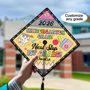 May include: A graduation cap with a yellow background and colorful graphics. The cap says "Kindergarten Grad! Next Stop 1st Grade!" with the year 2026. The name "Amelia" is also displayed. The cap is held up against a blurred outdoor background.