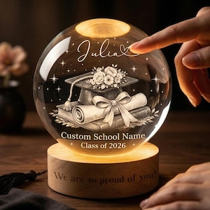 May include: A clear glass graduation sphere with a wooden base. Inside the sphere is a graduation cap, diploma, and books. The sphere is lit from within. The base has the text "We are so proud of you!" and the sphere has the name "Julia".
