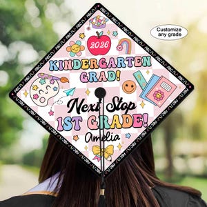 May include: A graduation cap with a colorful design. The cap features the text "Kindergarten Grad! Next Stop 1st Grade!" along with the name "Amelia" and the year "2026". The design includes illustrations of an apple, rainbow, and school supplies.