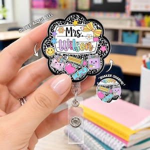 May include: A flower-shaped shaker badge reel featuring a black and white composition notebook pattern background. The center displays the text Mrs. Wilson with colorful school supply graphics including a pencil, apple, crayons, and a smiley face.